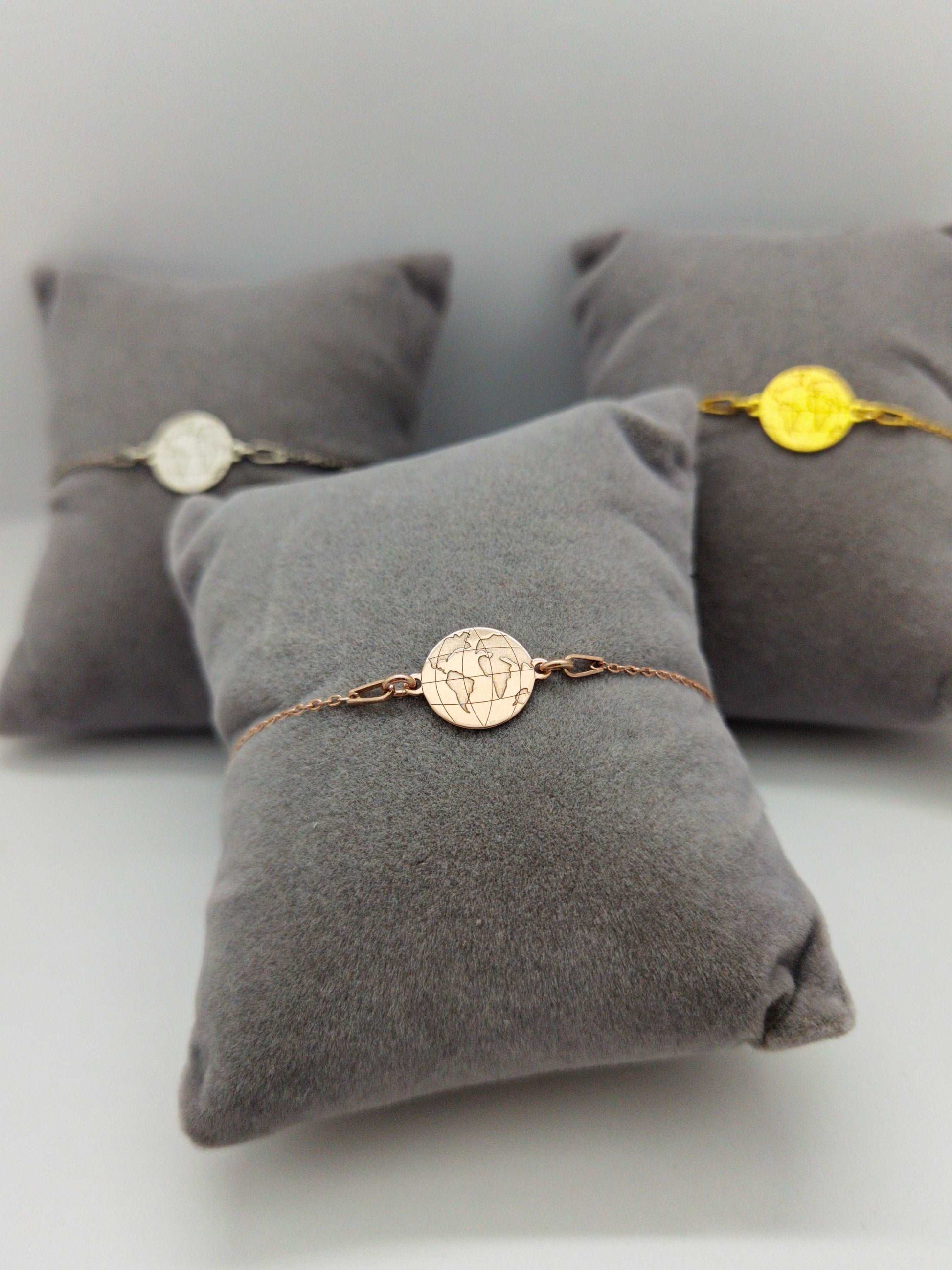 Drei graue Schmuckkissen mit Weltkarten Armbändern in silber, gold und roségold. Roségoldenes Armband im Fokus