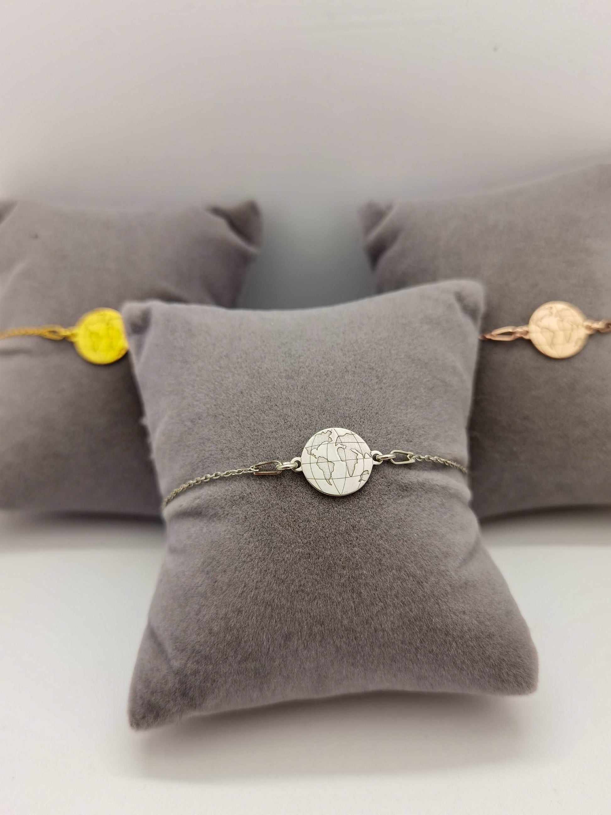 Drei graue Schmuckkissen mit Weltkarten Armbändern in silber, gold und roségold. Silbernes Armband im Fokus